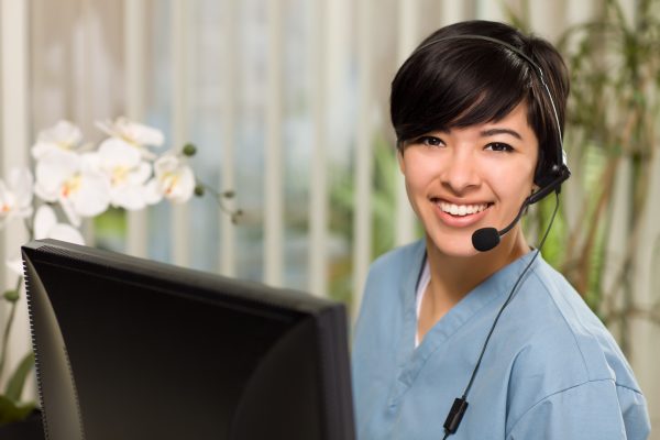 Smiling Attractive Multi-ethnic Young Woman Wearing Headset and Scrubs Near Her Computer Monitor.