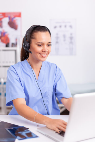 Medical receptionist wearing headset with microphone in private hospital typing on laptop Health care physician sitting at desk using computer in modern clinic looking at monitor